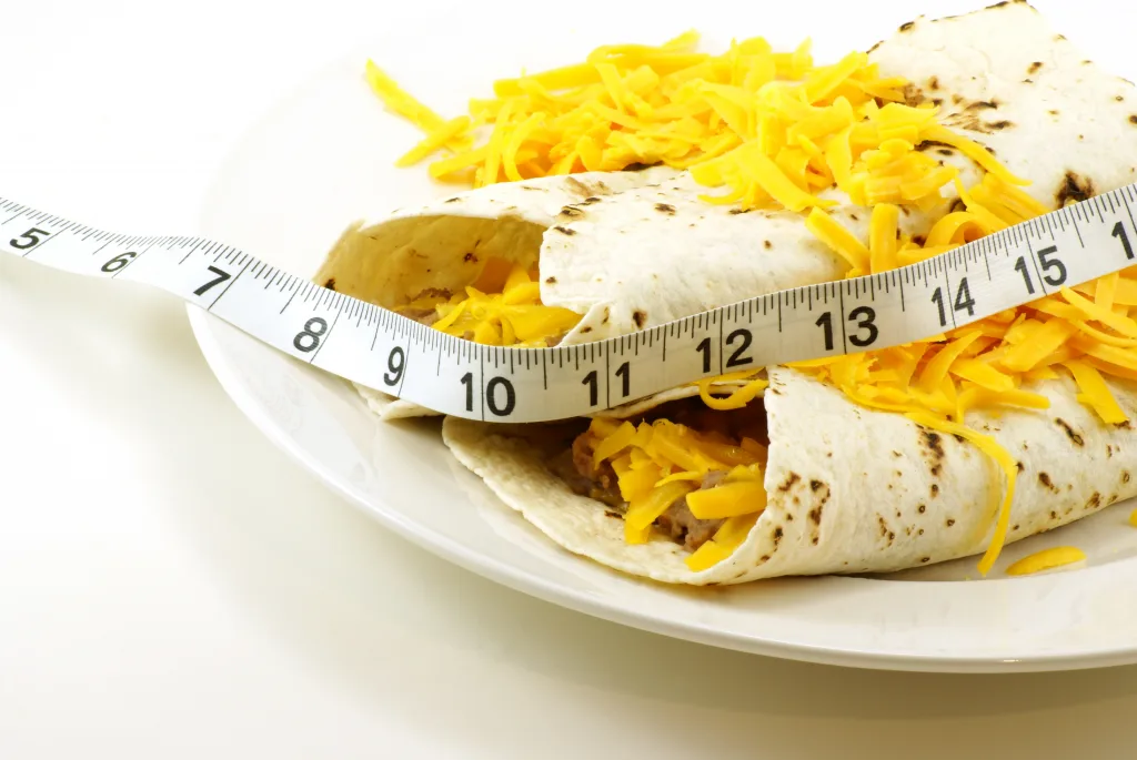 Homemade beef and bean mexican/spanish food burritos wrapped in grilled white tortillas with shredded cheddar cheese. Measuring tape draped across for fitness and weight loss concepts.