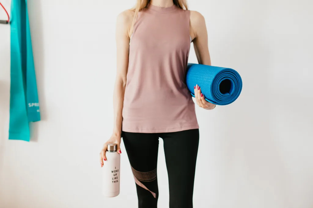 Standing woman holding yoga mat and tumbler for before exercise for weight loss