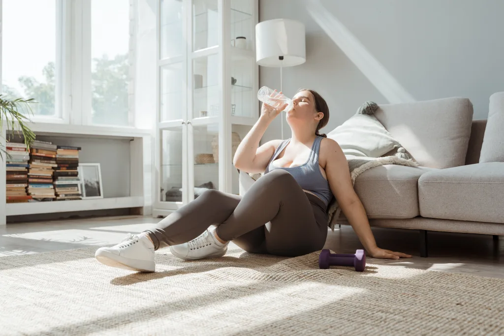 Tired overweight woman sitting on the floor drinking water after work out