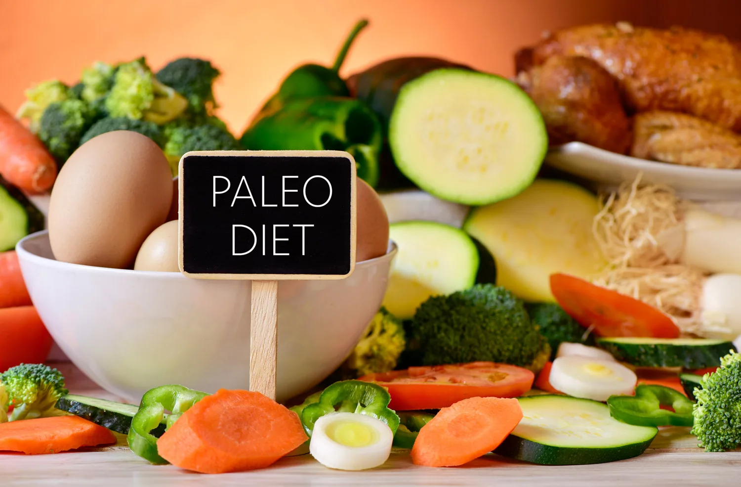 closeup of a signboard with the text paleo diet on a table full of different raw vegetables, a bowl with some chicken eggs and a chicken raw diet